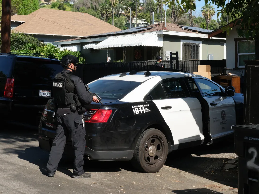 A police officer in tactical gear stands next to an LAPD patrol car.