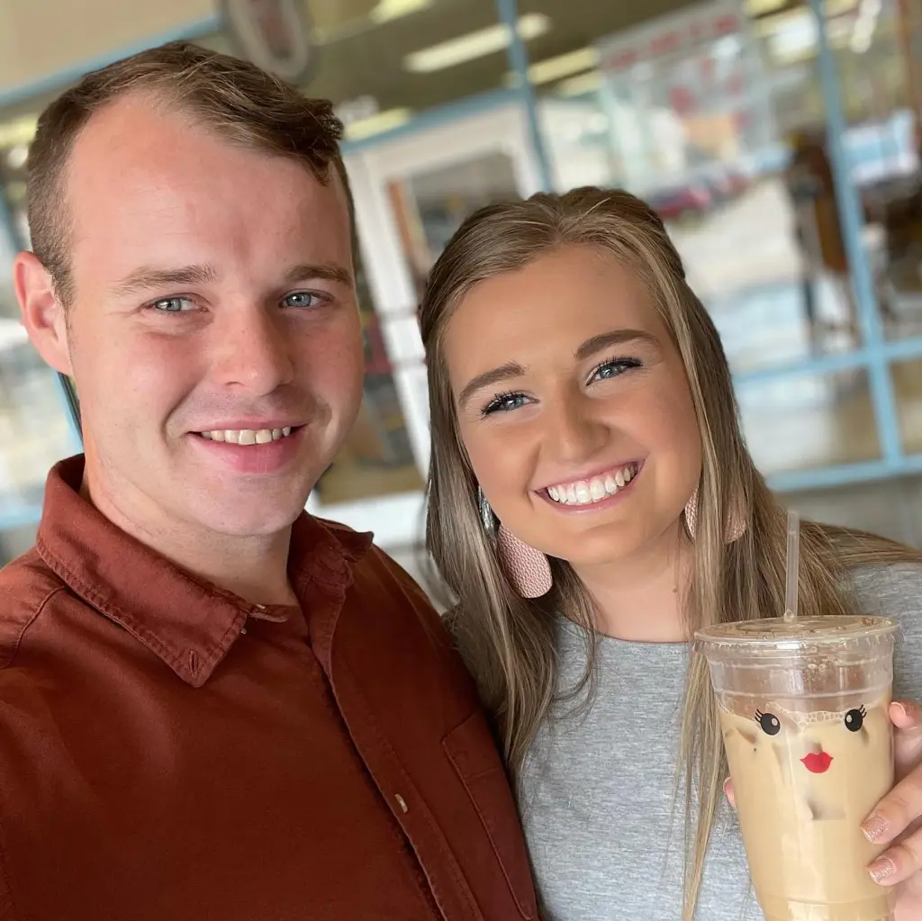 Joseph and Kendra Duggar smiling with Kendra holding an iced coffee with a face sticker on it.