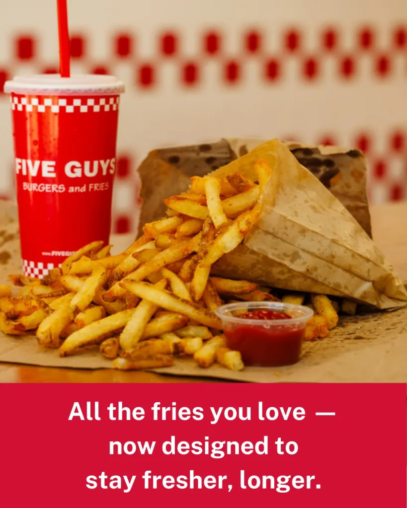 Five Guys fries spilling from a paper bag next to a cup of ketchup and a Five Guys drink cup.