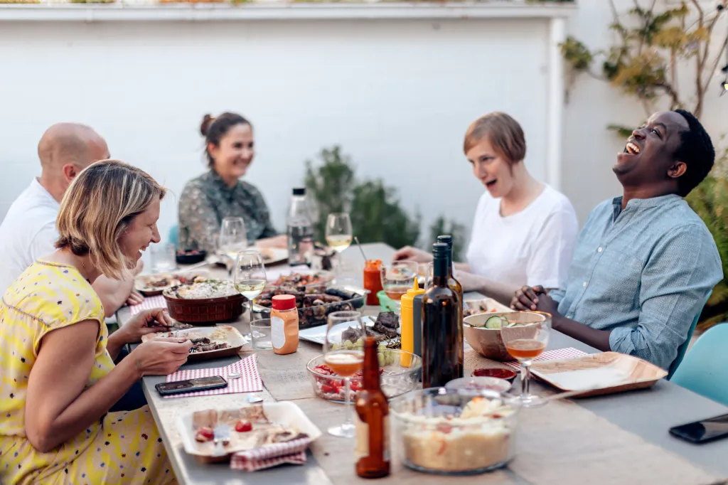 Multiracial friends eating and cheering together at an outdoor terrace table, with focus on the left woman's face.