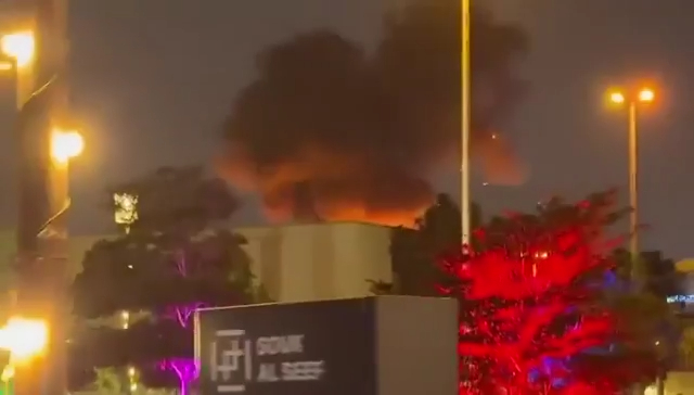Large plume of smoke and flames rising from a building at night, with streetlights and trees in the foreground.