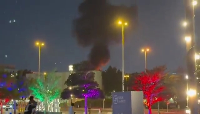 Thick black smoke rises into the night sky above buildings.