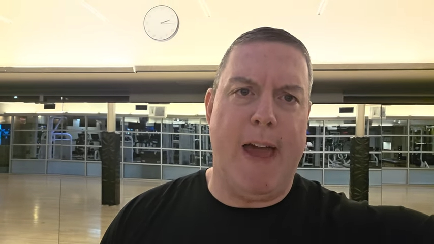 Samuel McVey, a former New Rochelle Spanish teacher, is seen speaking in front of a gym with exercise equipment visible through glass windows in the background.