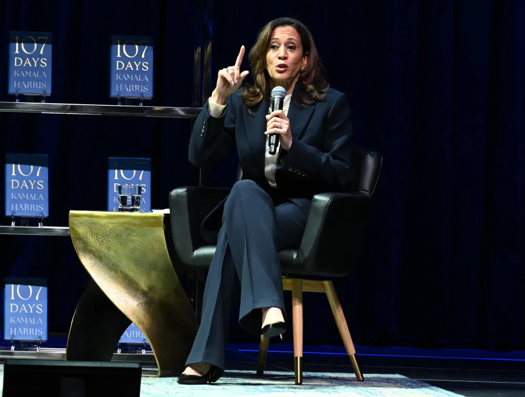 Kamala Harris speaking into a microphone on stage, with copies of her book 