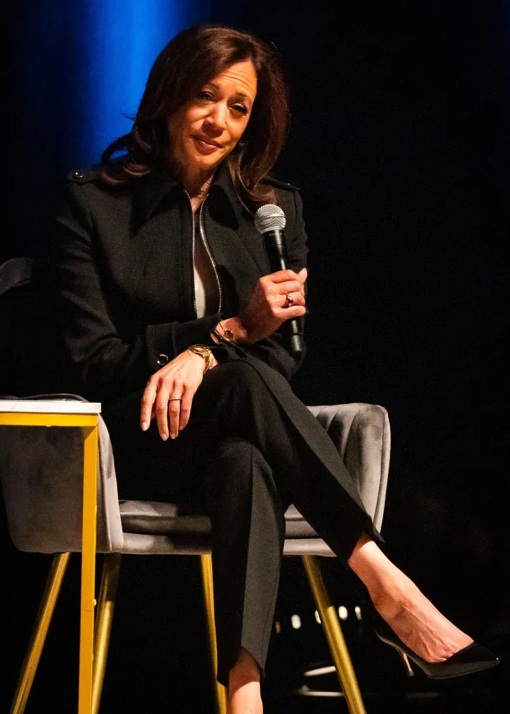 Kamala Harris, wearing a black pantsuit, sits holding a microphone while speaking at her