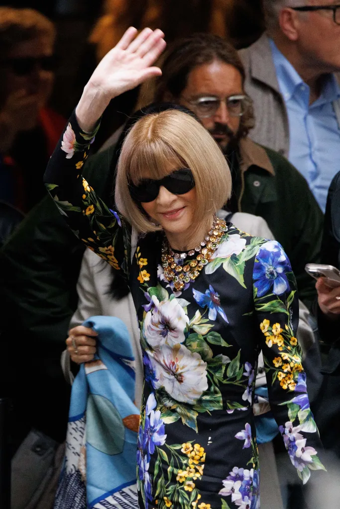 Anna Wintour in a black floral dress and dark sunglasses, waving to the crowd.