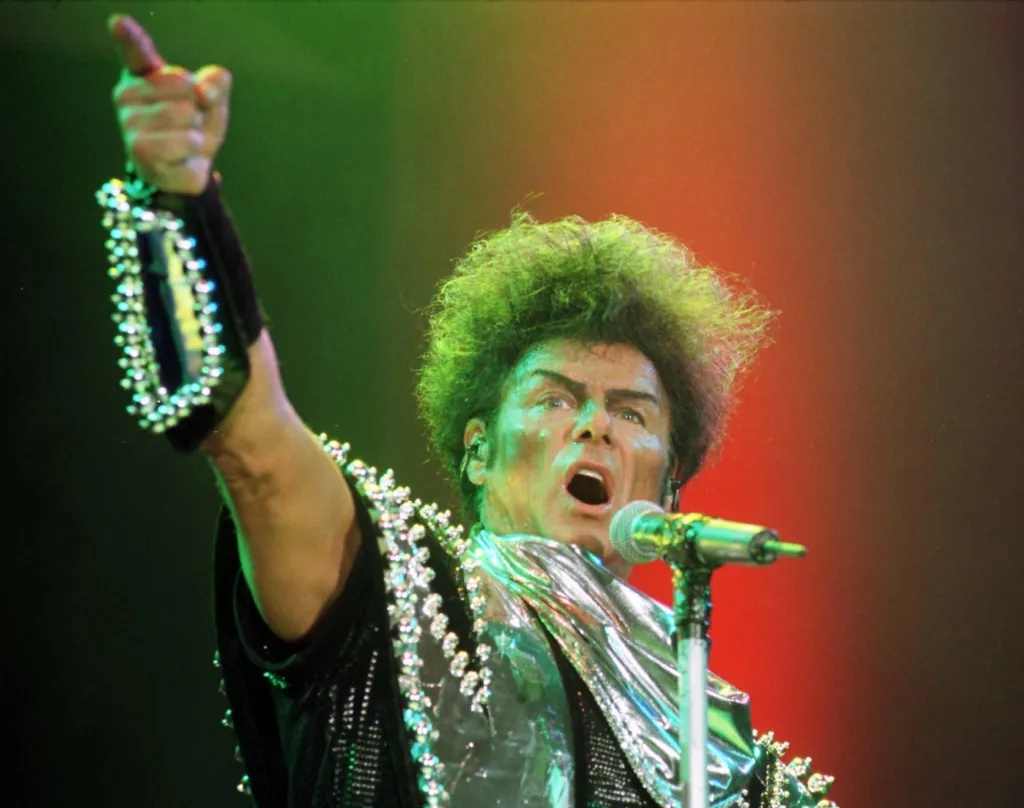 Pop star Gary Glitter performing on stage, dressed in a sparkling costume, with his arm raised and pointing.
