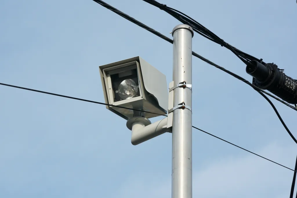 A speed camera mounted on a pole against a clear blue sky, with wires crisscrossing in the foreground and background.