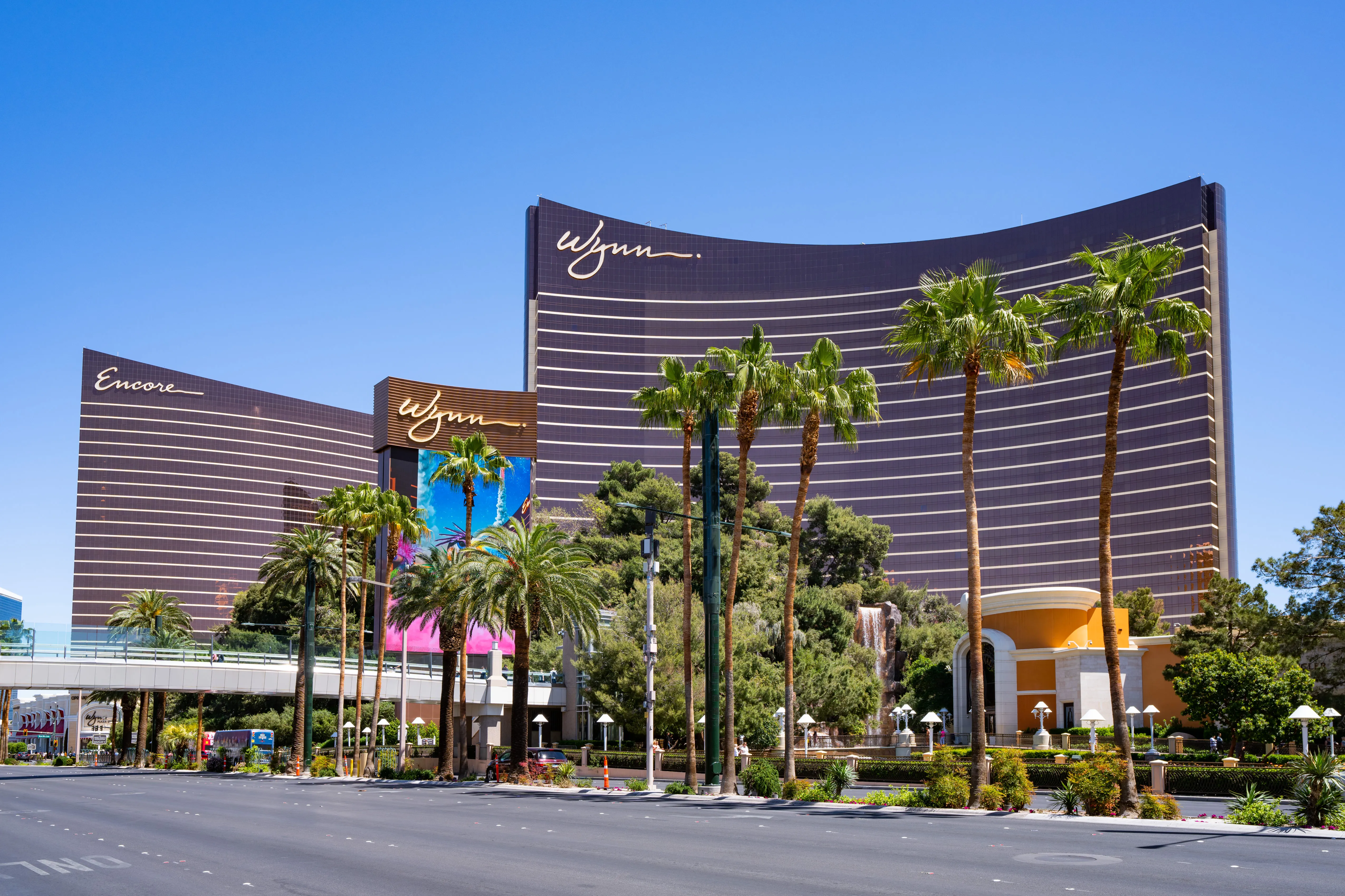 The Wynn Las Vegas and Encore Las Vegas hotels stand against a clear blue sky, with palm trees and landscaping in the foreground.