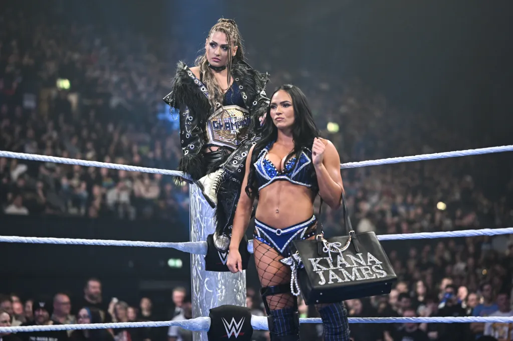 Giulia James, wearing a championship belt, stands on the turnbuckle with Kiana James beside her holding a handbag with her name on it.