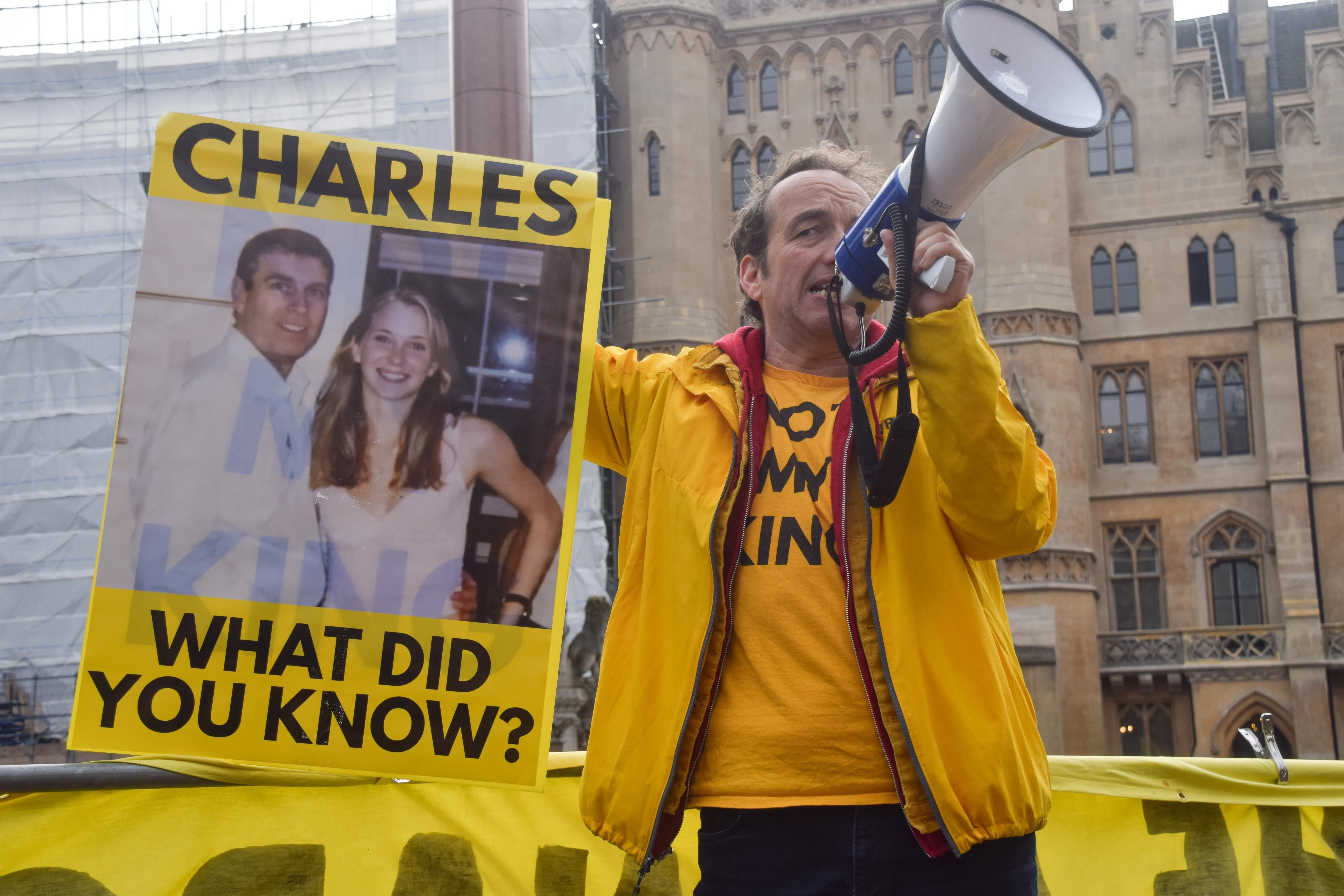 A man in a yellow jacket uses a megaphone and holds a protest sign featuring a photo of Andrew Mountbatten-Windsor and Virginia Giuffre with the text 