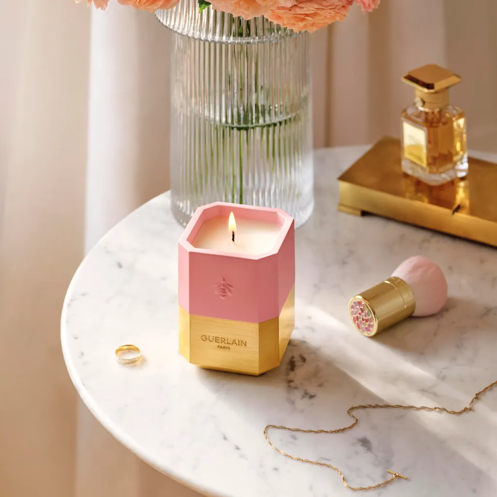 A Guerlain L'Art de Vivre Collection Scented Candle in a pink and gold hexagonal holder, lit on a marble table with a gold ring, gold necklace, perfume bottle, and makeup brush.