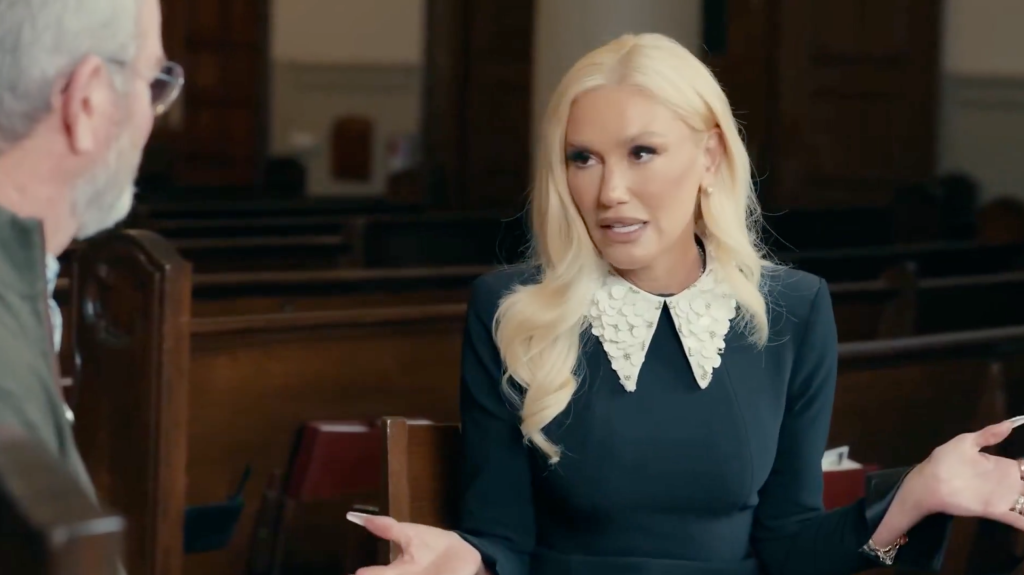Gwen Stefani in a dark dress with a white collar, gesturing with both hands as she speaks to someone off-camera.