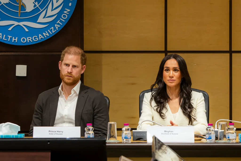 Prince Harry and Meghan Markle sitting at a table with their name cards in front of them, beneath a World Health Organization logo.