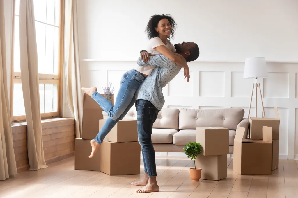 An African-American man holding his wife in his arms in their new home surrounded by moving boxes.