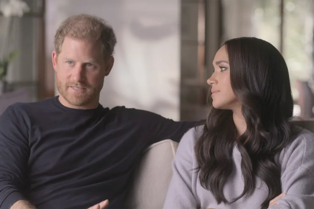 Prince Harry, in a black top, looks towards Meghan Markle, in grey sweater, as they sit together on a couch for their Netflix show.