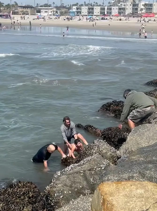 People helping two girls out of the water on a rocky shore.