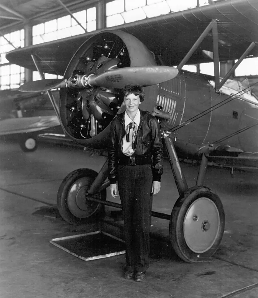 Amelia Earhart stands in front of an airplane.
