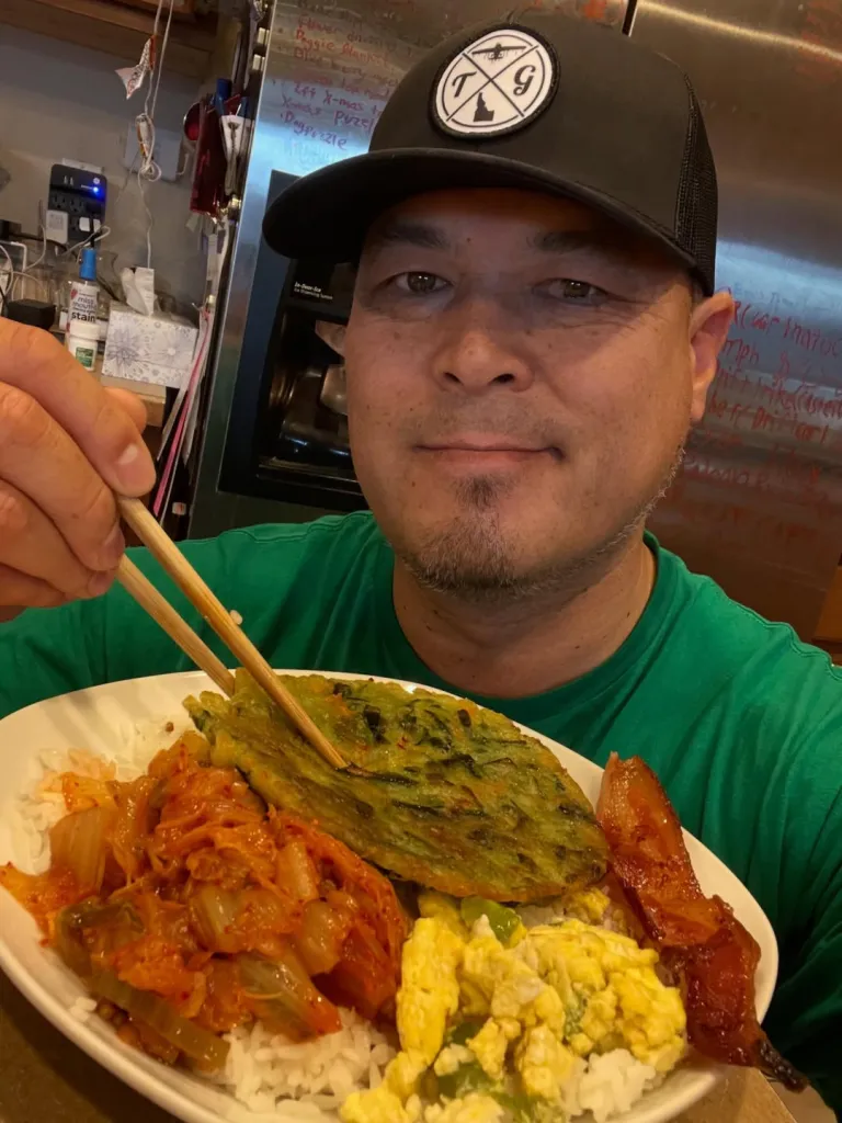 A man in a green shirt and black cap holds chopsticks over a plate of kimchi, rice, a savory pancake, scrambled eggs, and bacon.