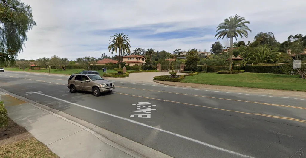 A beige SUV drives down El Apajo Road past a large residential property with palm trees and a gated entrance.