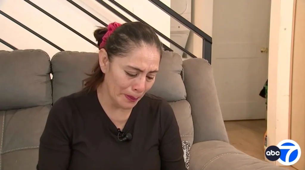 A grieving mother sits on a sofa, looking downward with tears in her eyes.