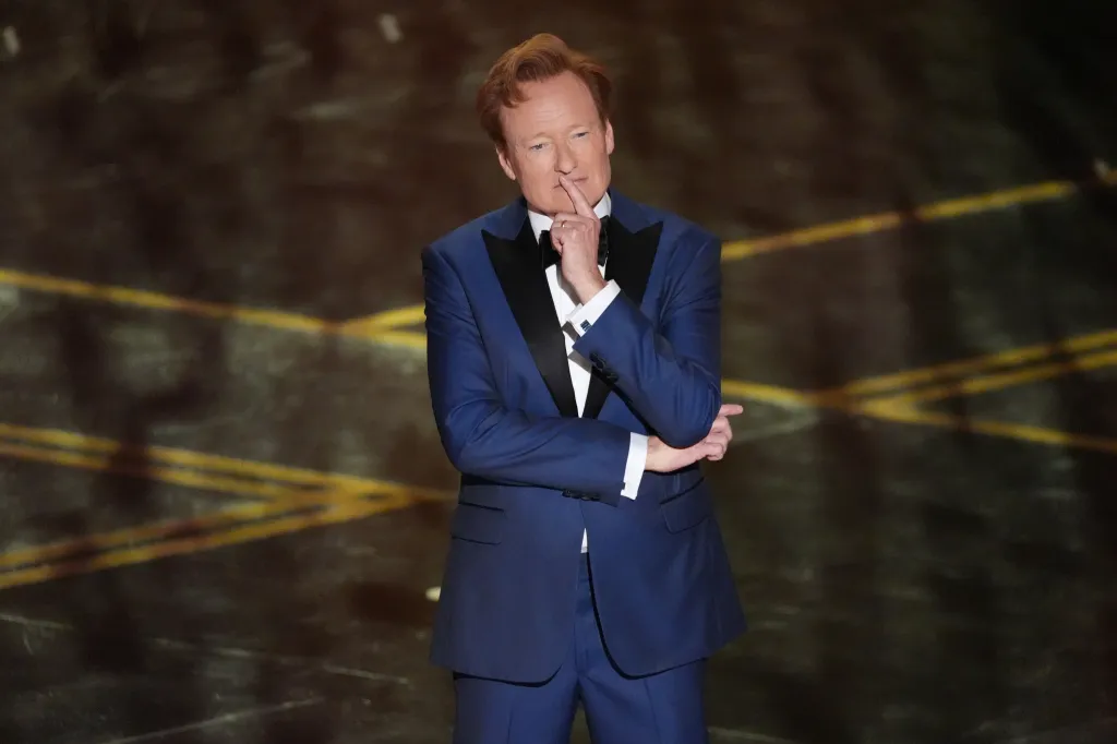 Conan O'Brien in a blue tuxedo with his finger to his mouth as if in thought.