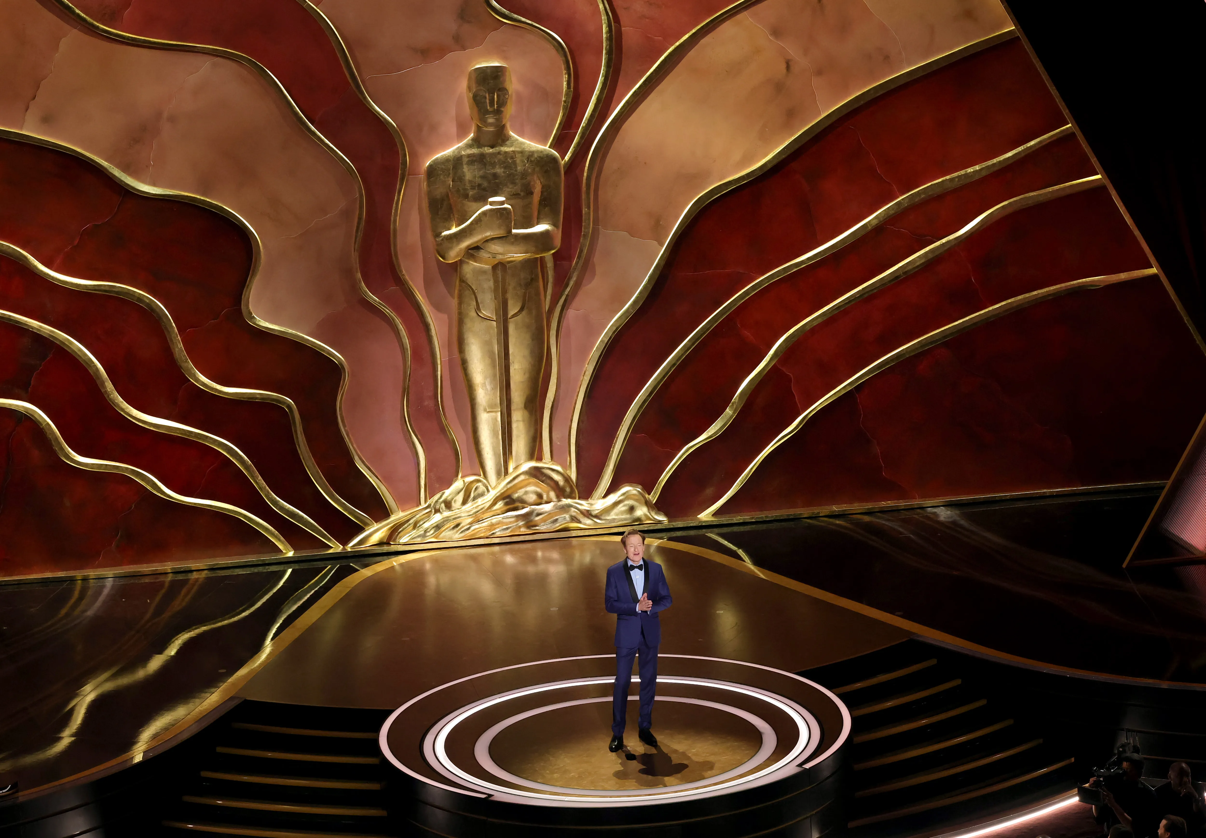 Conan O'Brien speaking onstage at the 97th Annual Oscars with a large golden Oscar statue behind him.