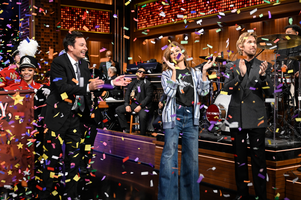 Jimmy Fallon, Eva Mendes, and Ryan Gosling clapping as confetti falls around them on a talk show set.