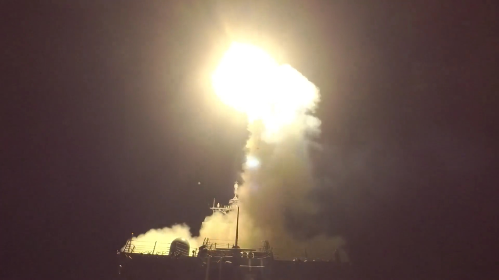 A missile launch from a ship at night, emitting a bright flare and smoke.