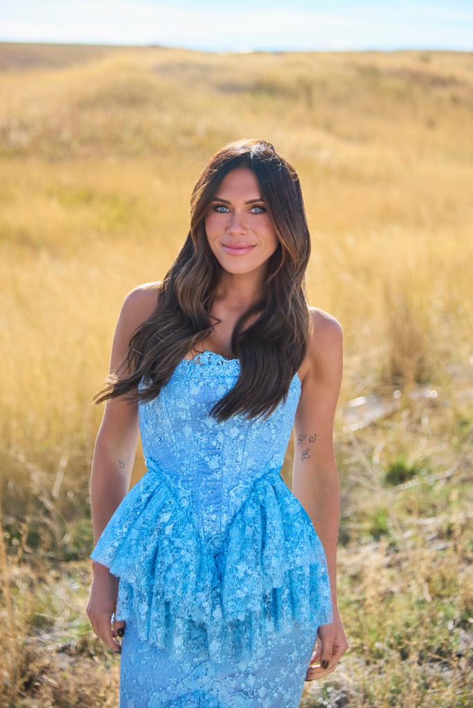 Jessi Ngatikaura smiling in a blue lace dress in a field of dry grass.