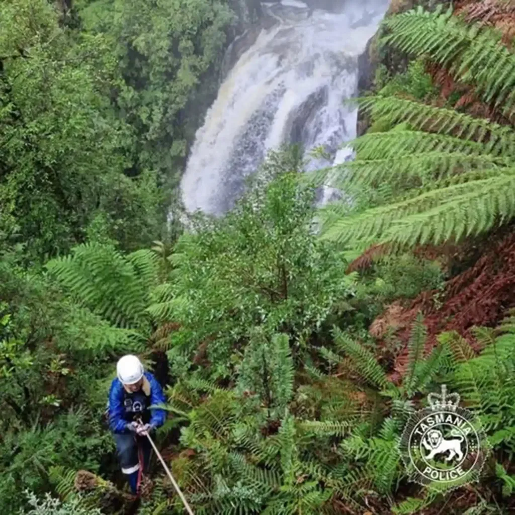 A person in a blue jacket and white helmet rappelling down a waterfall in a lush, green forest.