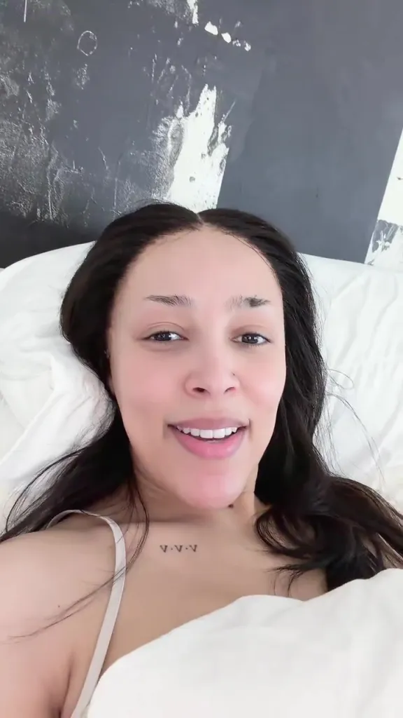 Doja Cat smiling while lying in bed, showing her tattoo.