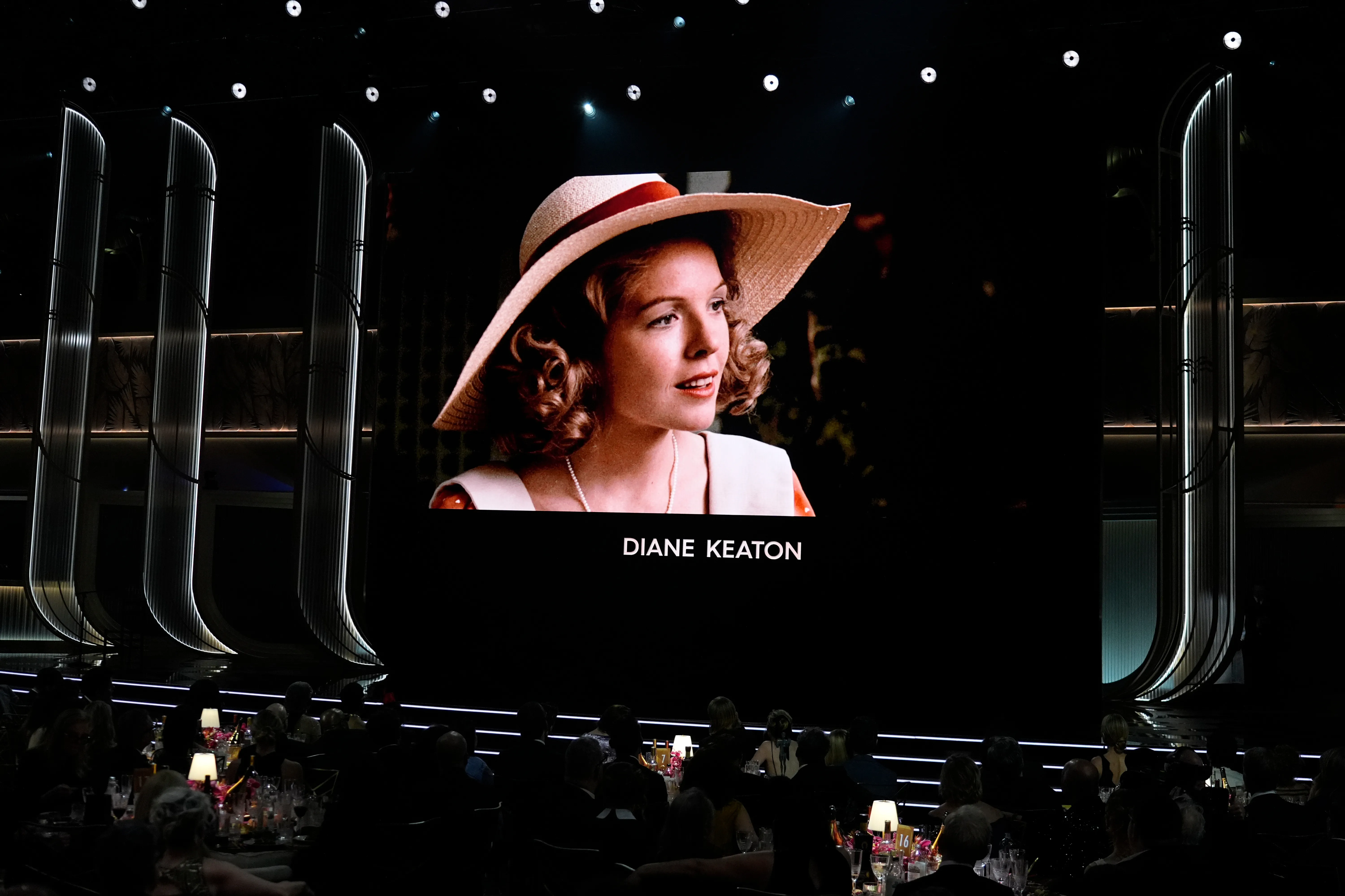 A large screen at an awards show displays a portrait of Diane Keaton wearing a straw hat with a red band, with her name beneath.