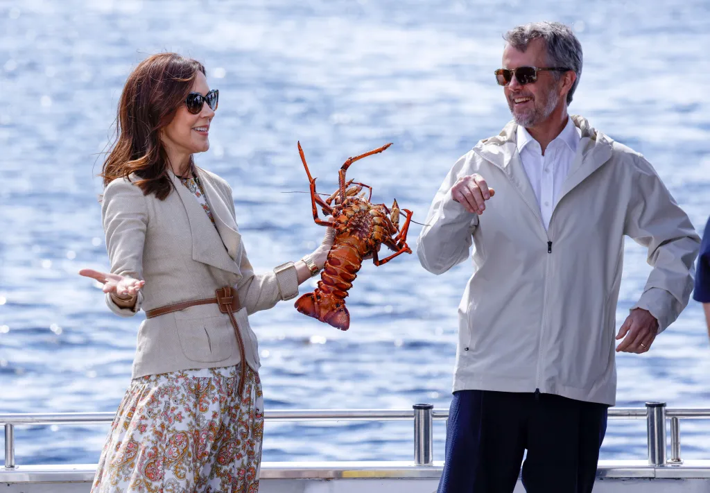 Queen Mary of Denmark holds up a large red rock lobster as King Frederik X of Denmark smiles beside her.