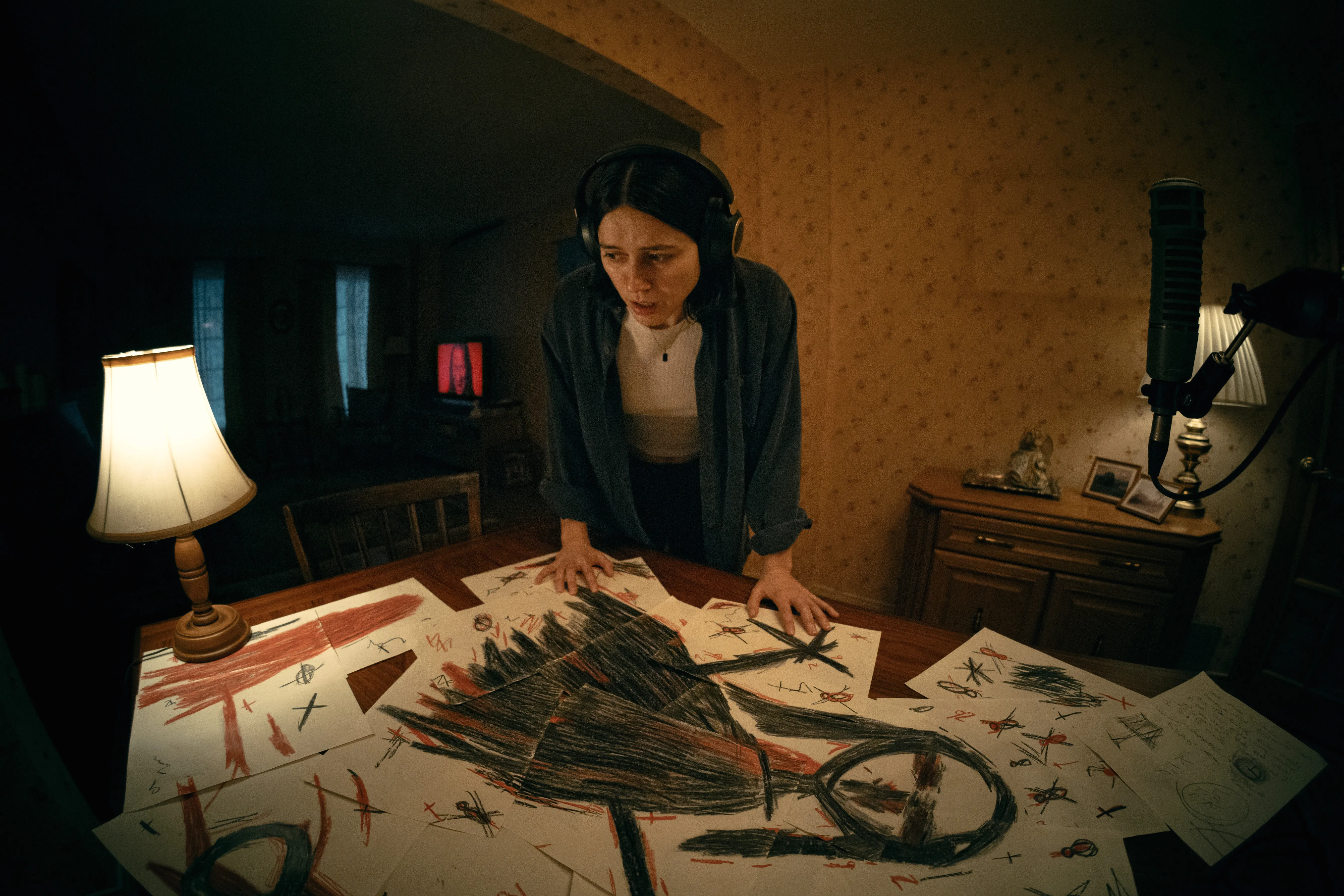Nina Kiri looks down at numerous disturbing crayon drawings scattered across a table in a scene from the horror movie 