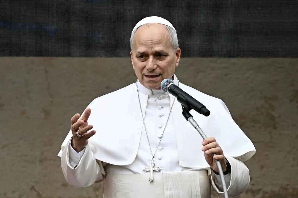 A Catholic bishop or cardinal in white robes speaks into a microphone.