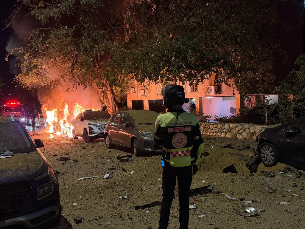 A first responder stands near burning vehicles and debris after a missile attack.