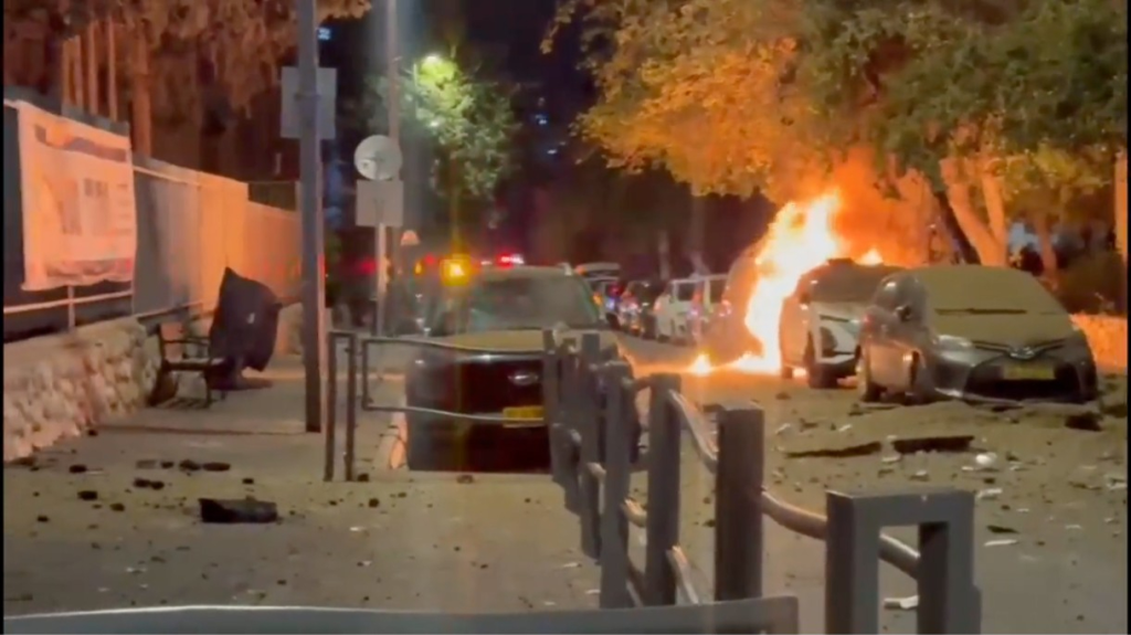 Two cars on fire in an urban street at night, with debris scattered on the ground.