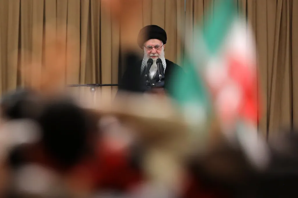 Iranian Supreme Leader Ali Khamenei speaking at a meeting, with a blurred Iranian flag in the foreground.