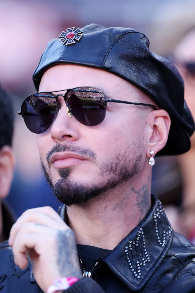 J Balvin, wearing a black leather beret and sunglasses, watches from the field.