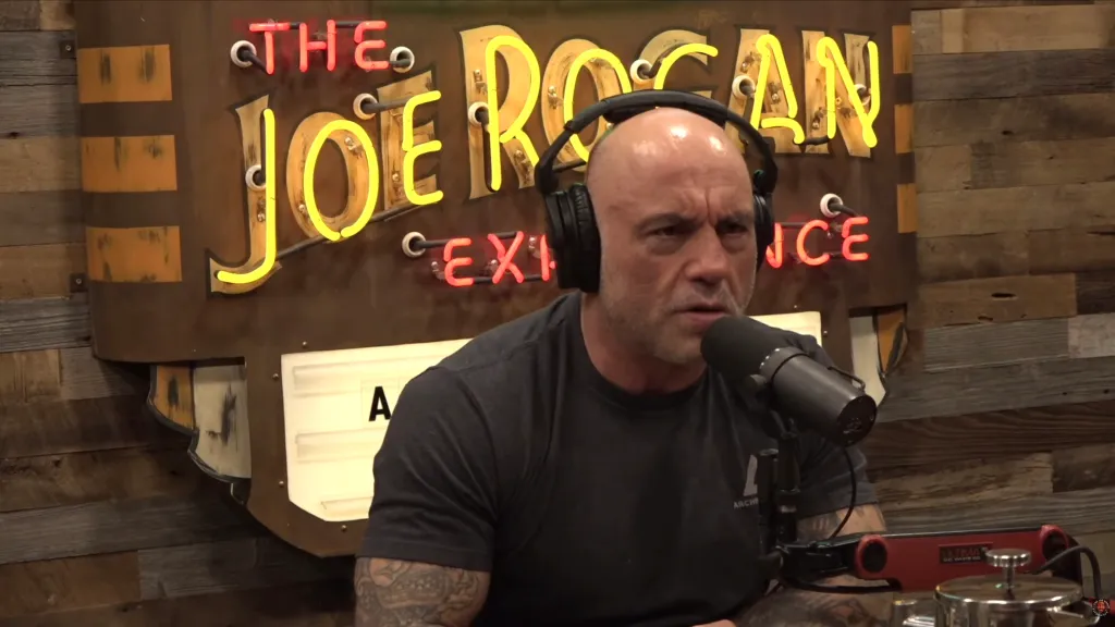 Joe Rogan speaking into a microphone with his podcast sign in the background.