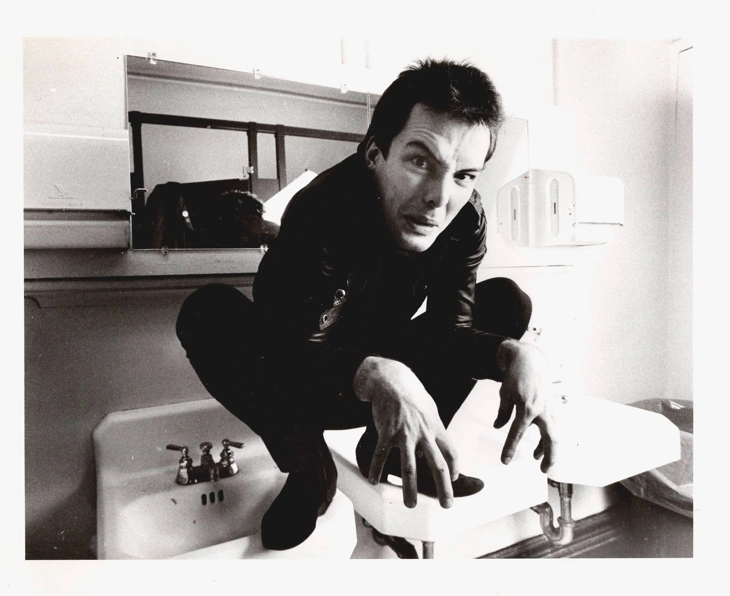 Jello Biafra in a black leather jacket squats on a bathroom sink, staring at the camera.