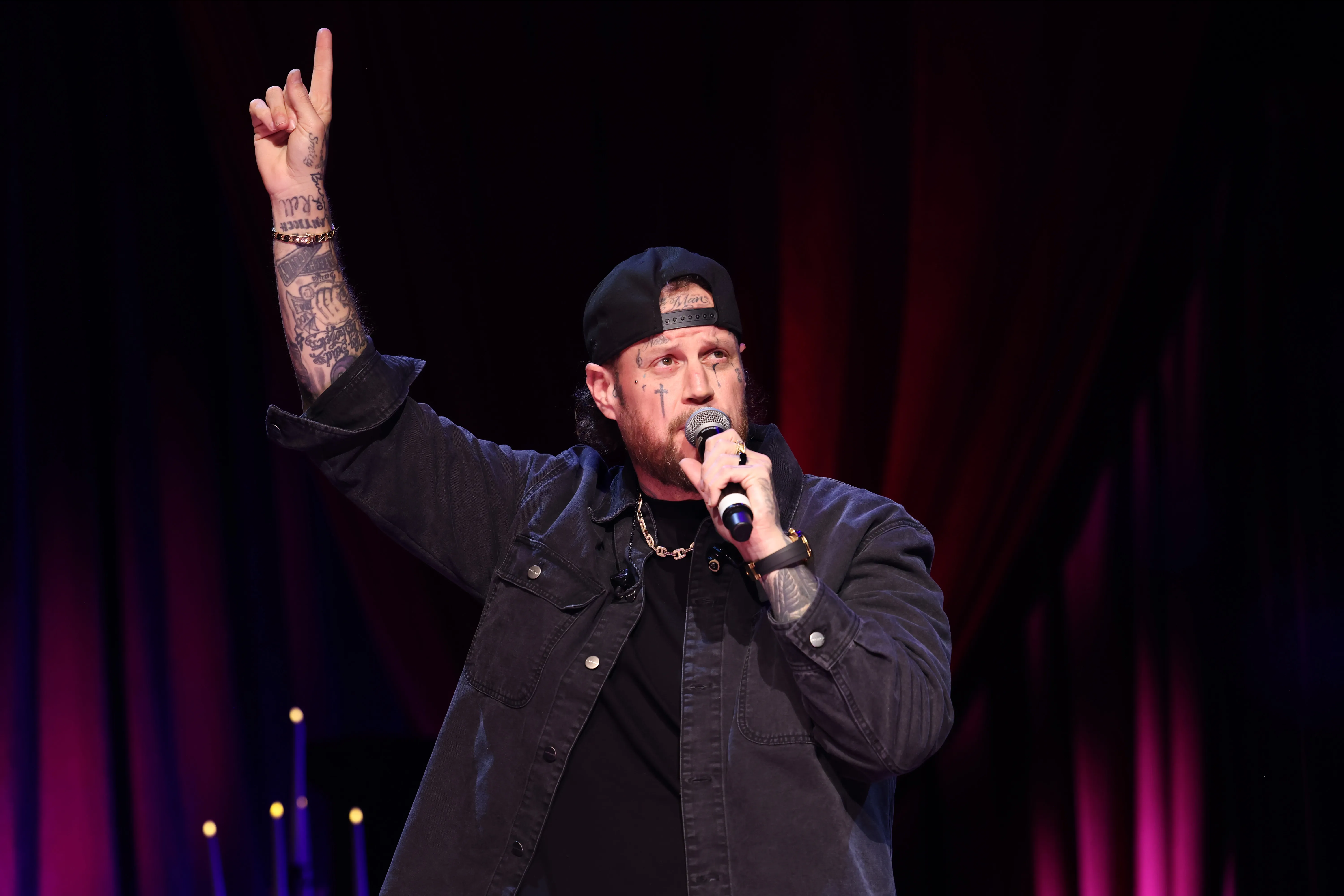 Jelly Roll performs onstage, holding a microphone and raising a tattooed arm with his index finger pointed up.