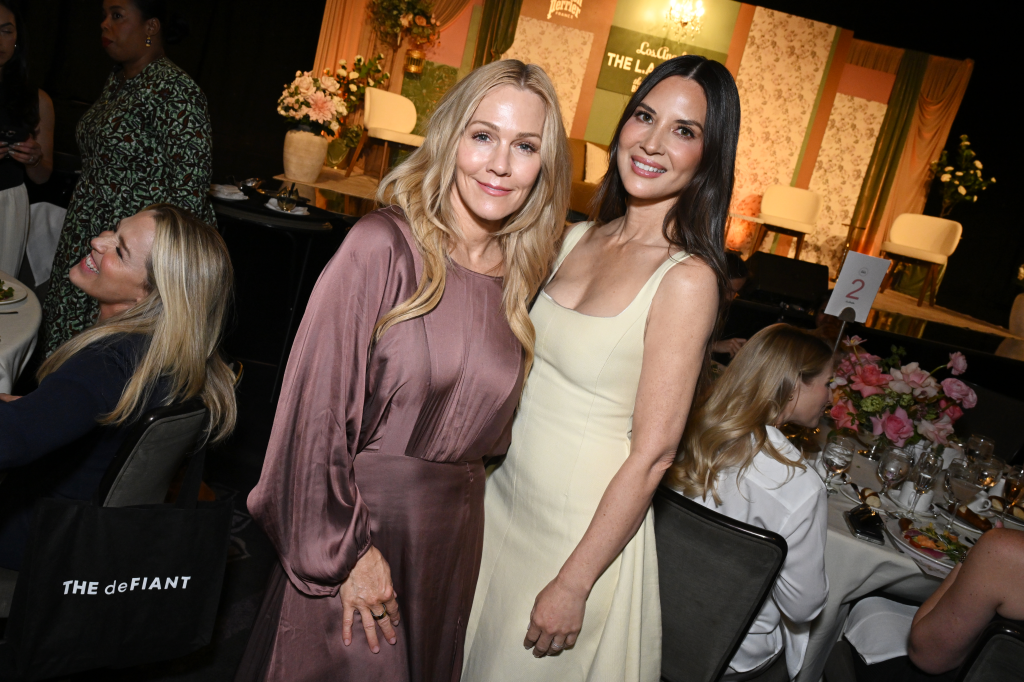Jennie Garth in a brown dress and Olivia Munn in a light yellow dress smiling at the LA Magazine Women Of Impact 2026 luncheon.
