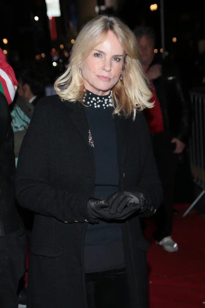 Jennifer Runyon smiling with blonde hair, wearing a black jacket and a black top with pearl details and a colorful brooch.