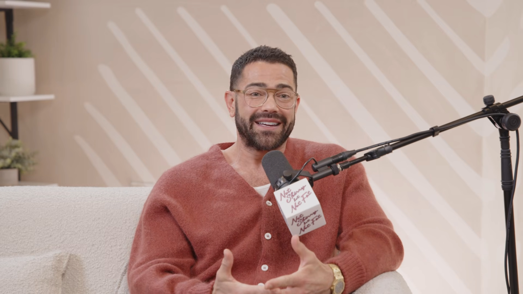 Jesse Metcalfe, wearing glasses and a reddish-brown cardigan, speaks into a microphone during an interview.