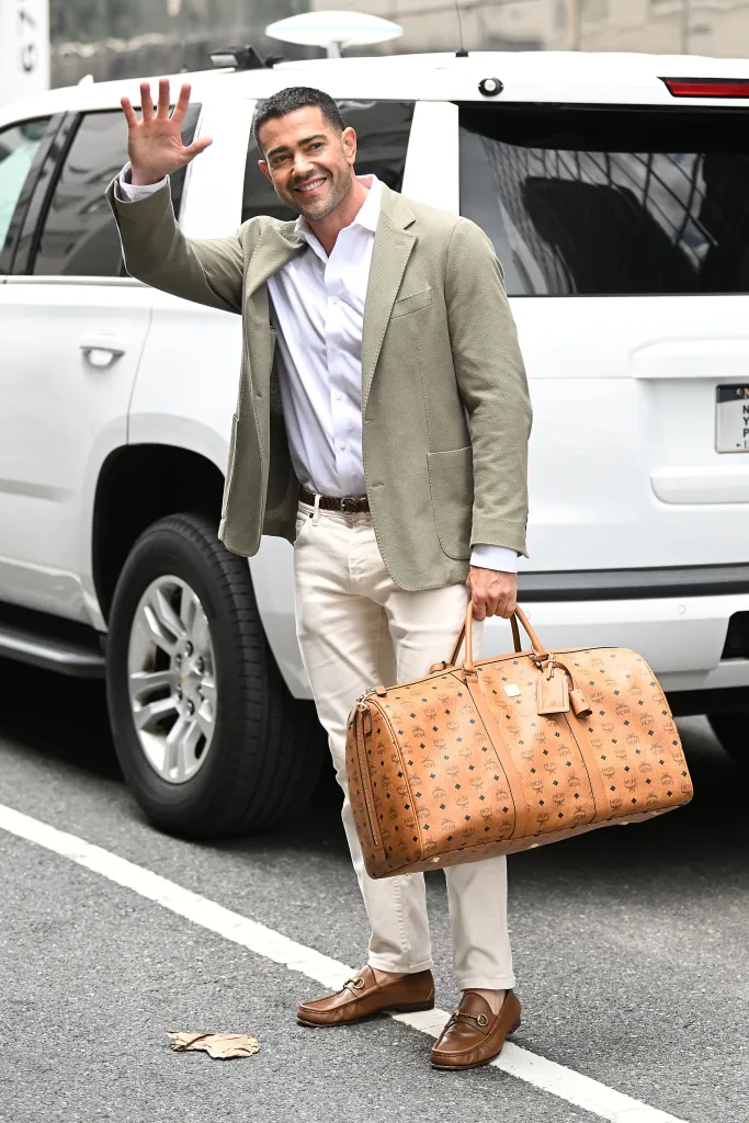 Jesse Metcalfe smiling and waving while holding a brown MCM duffel bag.