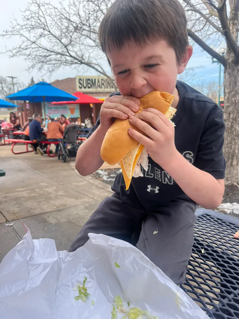 Young boy eating a submarine sandwich outdoors.