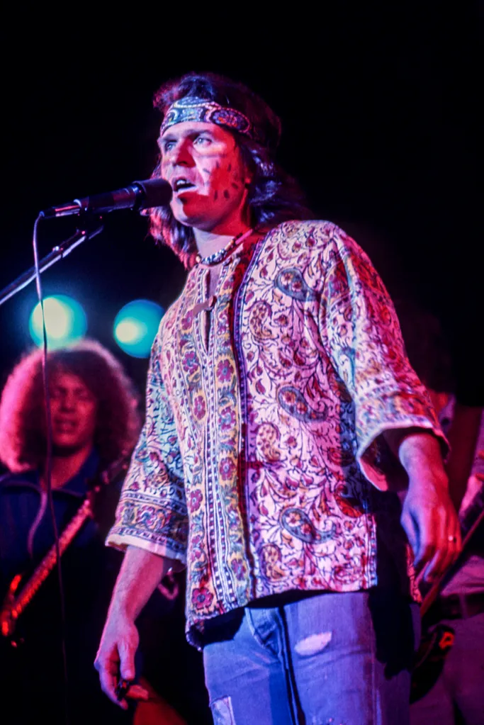 Joe McDonald performing live with a microphone, wearing a patterned shirt and bandana.