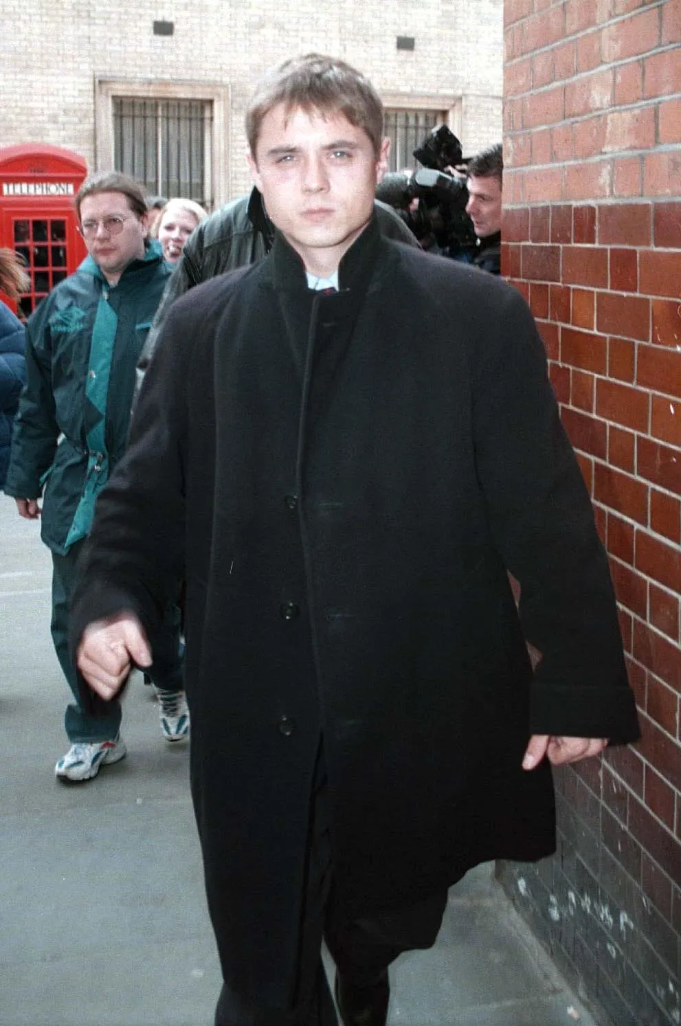 John Alford in a black coat, with people behind him, walking past a brick wall and a red telephone box.